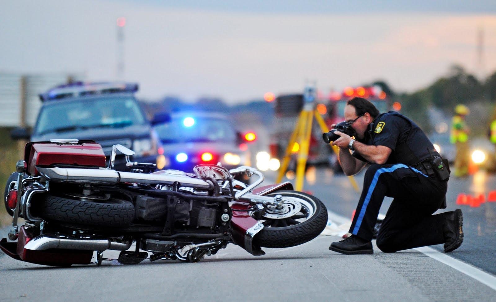 motorcycle accident scene with emergency vehicles in the background and a police officer kneeling in the foreground taking a photo of the toppled motorcycle