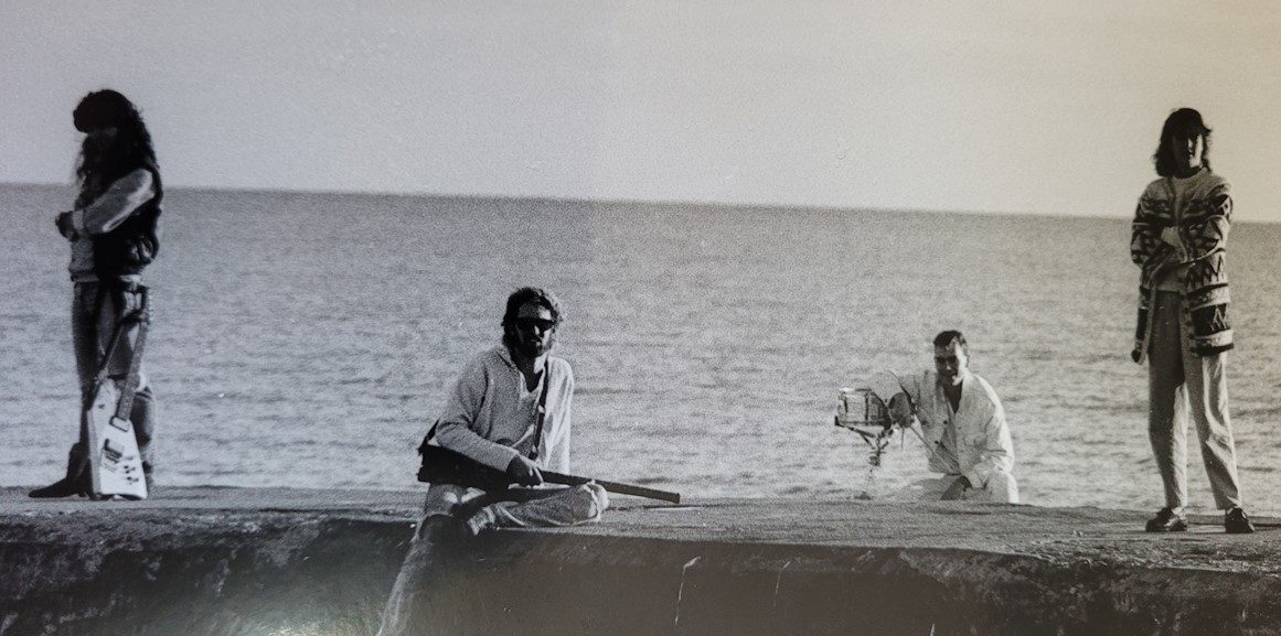 b&w of four musicians sitting on a pier with a lake in the background